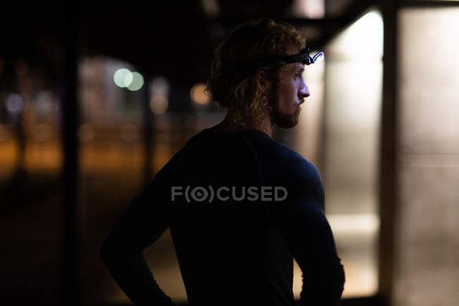 Side view of a young athletic Caucasian man exercising in a city park in the evening, with a headlight on resting during a break with defocused lights and buildings in the background — Stock Photo
