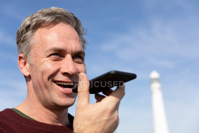 Front view close up of a middle-aged caucasian man talking on a smartphone while enjoying free time relaxing on a beach near a lighthouse on a sunny day — Stock Photo