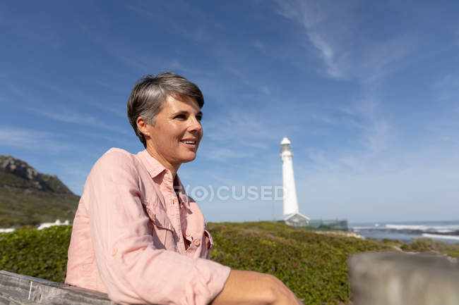Vista lateral de cerca de una mujer caucásica de mediana edad disfrutando de tiempo libre sentada en un banco relajándose en una playa cerca de un faro al lado del mar en un día soleado - foto de stock