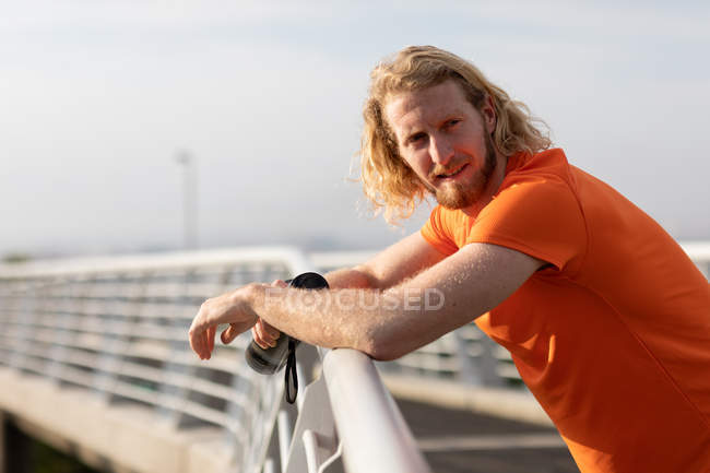 Retrato de um jovem atlético caucasiano exercitando em uma passarela em uma cidade, apoiando-se no corrimão e olhando para a câmera — Fotografia de Stock