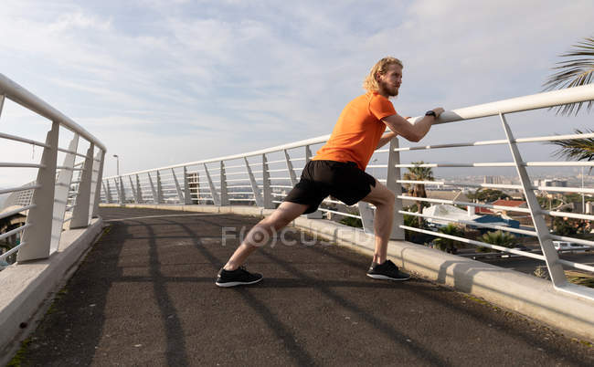 Seitenansicht eines jungen athletischen kaukasischen Mannes, der auf einer Fußgängerbrücke in einer Stadt trainiert, sich dehnt und am Geländer lehnt — Stockfoto