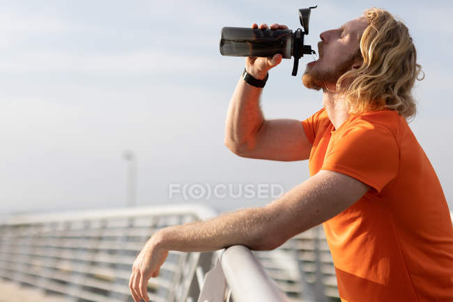 Side view of a young athletic Caucasian man exercising on a footbridge in a city, drinking water during a break — Stock Photo