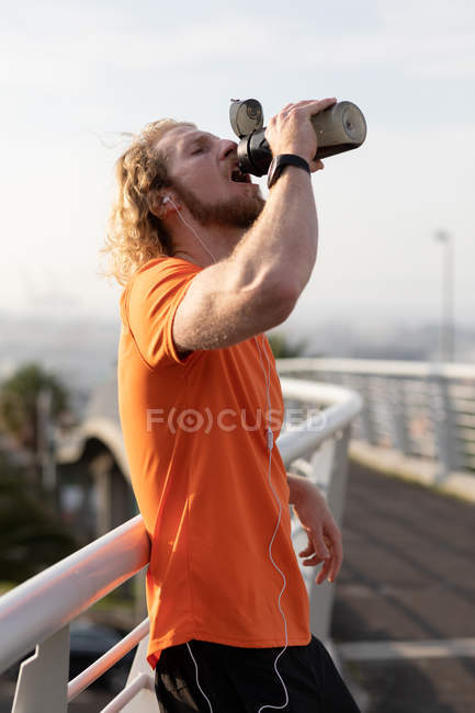 Side view of a young athletic Caucasian man exercising on a footbridge in a city, listening to music with earphones on drinking water during a break — Stock Photo