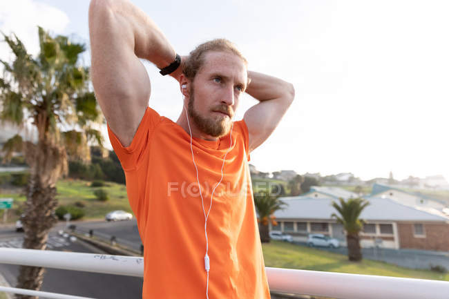 Side view of of a young athletic Caucasian man exercising on a footbridge in a city, listening to music with earphones on and stretching with his hands behind his head — Stock Photo
