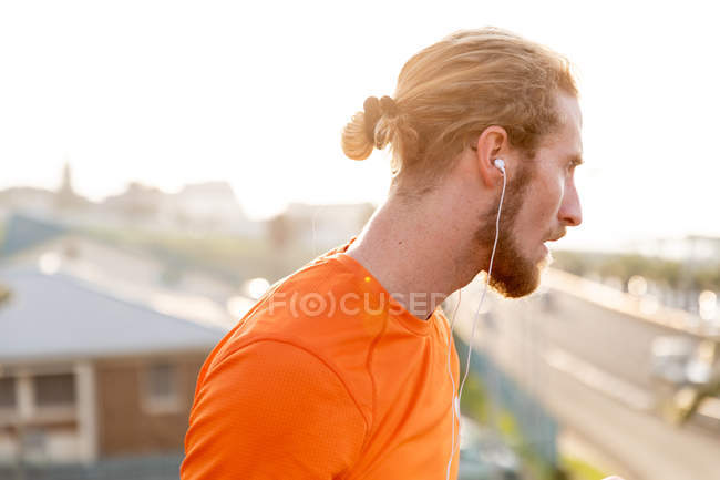 Side view of a young Caucasian man exercising on a footbridge in a city, listening to music with earphones on during a break — Stock Photo