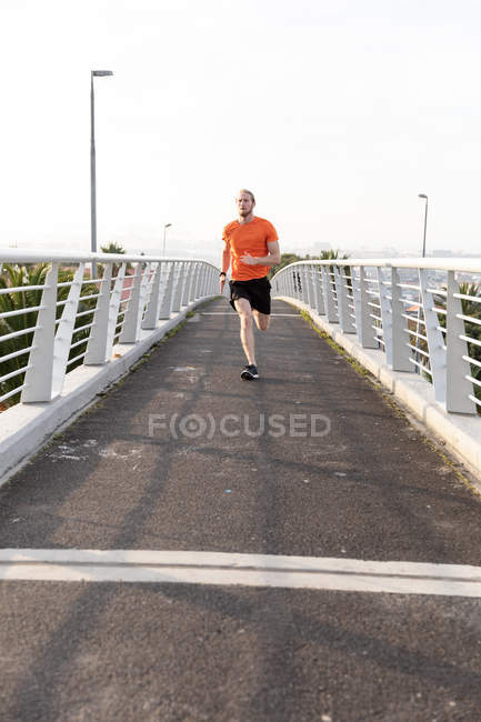 Передній вигляд молодого атлетичного кавказького чоловіка, який тренувався на пішохідному мосту в місті, бігав і слухав музику з навушниками. — стокове фото