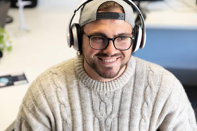 Vista frontal de un joven caucásico trabajando en un escritorio en una oficina creativa, usando gafas, gorra y escuchando música con auriculares, sonriendo - foto de stock