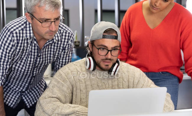 Frontansicht eines jungen kaukasischen Mannes, der an einem Schreibtisch in einem kreativen Büro arbeitet, während hinter ihm männliche und weibliche Kollegen stehen und auf seinen Bildschirm schauen. — Stockfoto