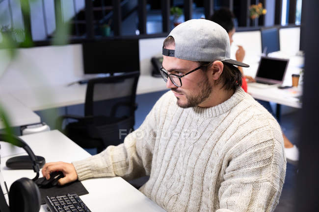 Side view of a young Caucasian man working at a desk using a computer in a creative office, wearing glasses and cap. — Stock Photo