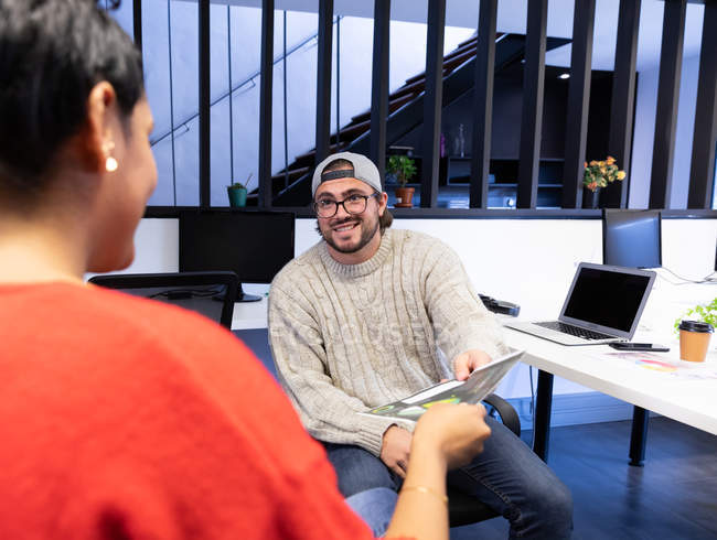 Front view of a young Caucasian man sitting at a desk in a creative office, talking to his female colleague, smiling, wearing glasses and cap. — Stock Photo