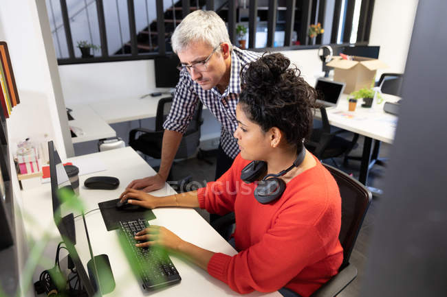 Seitenansicht einer jungen Frau mit gemischter Rasse, die an einem Schreibtisch in einem kreativen Büro arbeitet und einen Computer mit einem männlichen kaukasischen Kollegen benutzt, der hinter ihr steht und auf ihren Bildschirm blickt. — Stockfoto