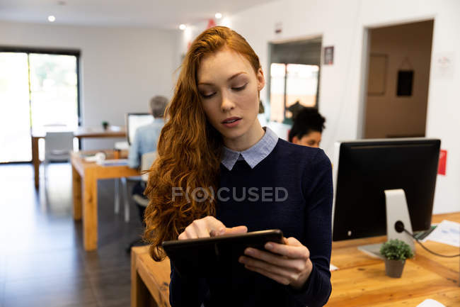 Vista frontal de una joven mujer caucásica que trabaja en una oficina creativa, de pie junto a su escritorio, sosteniendo una tableta con sus colegas en el fondo . - foto de stock