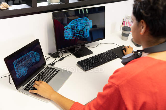 Вид сзади на молодую расистку, работающую за столом в креативном офисе, с помощью двух компьютеров, смотрящую на экраны с наушниками на шее . — стоковое фото