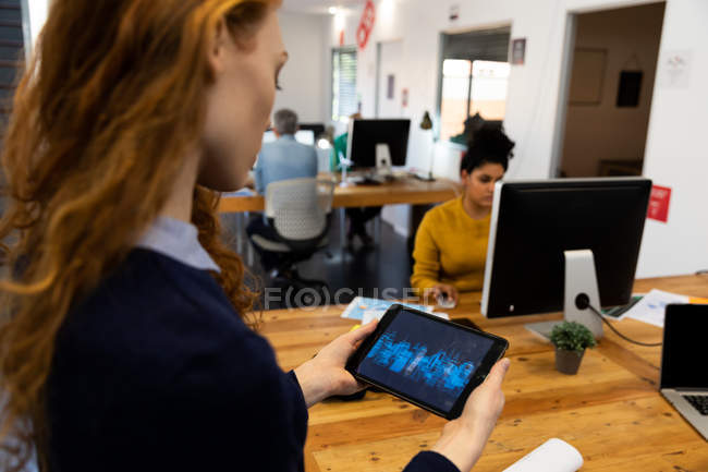 Вид сбоку: молодая кавказка, работающая в креативном офисе, стоящая у стола, держащая планшетный компьютер со своими коллегами на заднем плане . — стоковое фото