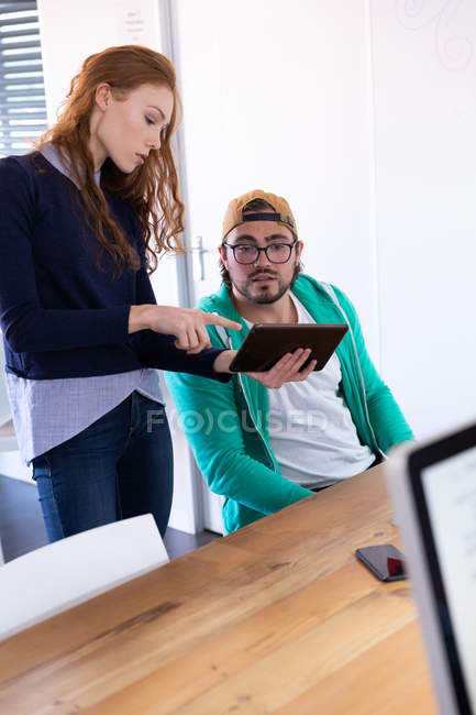 Вид сбоку молодой кавказки и мужчины, работающего в креативном офисе, женщины, стоящей у стола, держащей планшетный компьютер и обсуждающей со своим коллегой-мужчиной
. — стоковое фото