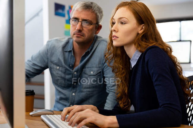 Бічний вид на молоду кавказьку жінку і кавказського чоловіка, який працює в офісі, дивиться на екран, жінка за допомогою комп'ютера, друкує на екрані. — стокове фото