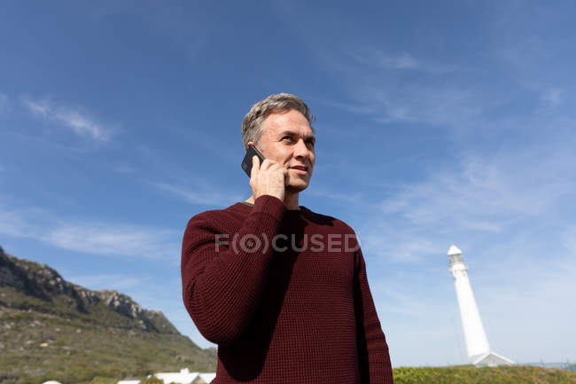 Front view of an adult Caucasian man talking on a smartphone standing on a beach near a lighthouse on a sunny day — Stock Photo