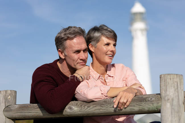 Vista frontal close-up de um casal adulto caucasiano desfrutando de tempo livre sentado em um banco perto de um farol em um dia ensolarado — Fotografia de Stock