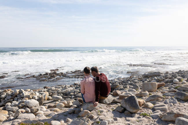 Visão traseira de um casal adulto caucasiano desfrutando de tempo livre relaxando juntos em uma praia ao lado do mar em um dia ensolarado — Fotografia de Stock