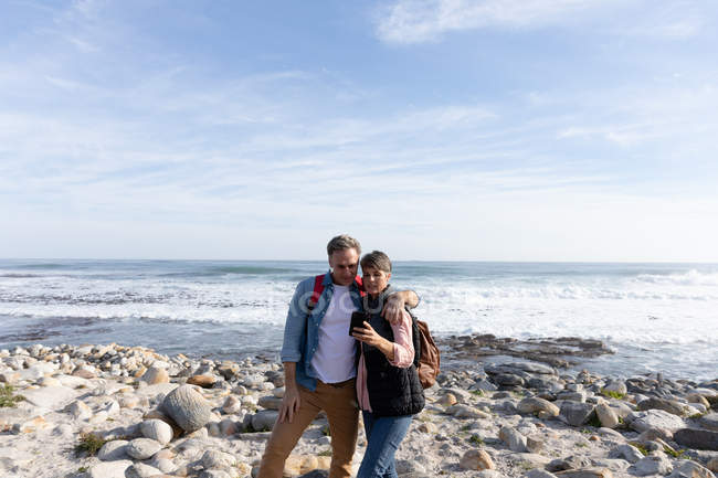 Vue de face d'un couple caucasien utilisant un smartphone tout en profitant de temps libre au bord de la mer sur une plage par une journée ensoleillée — Photo de stock