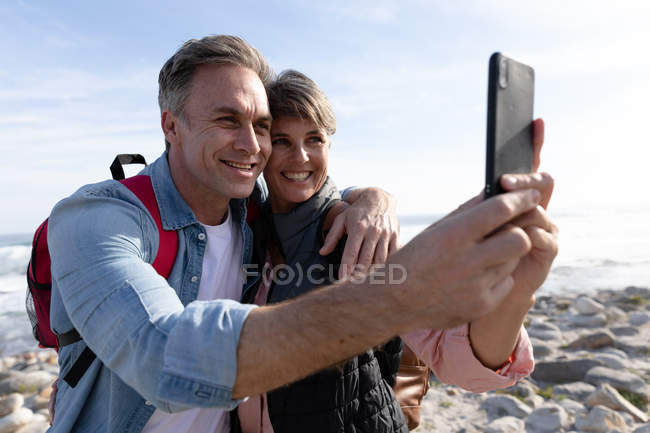 Vue de face gros plan d'un couple caucasien utilisant un smartphone tout en profitant de temps libre au bord de la mer sur une plage par une journée ensoleillée — Photo de stock