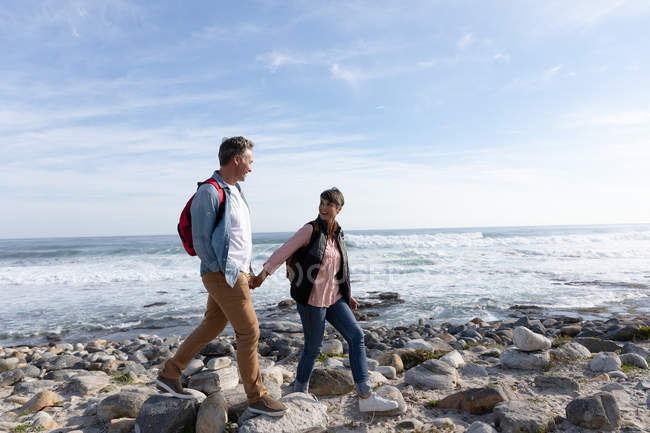 Vista laterale di una coppia caucasica adulta che si gode il tempo libero passeggiando su una spiaggia e sorridendo accanto al mare in una giornata di sole — Foto stock
