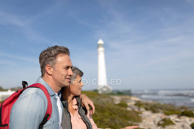 Vue de côté gros plan d'un couple adulte caucasien jouissant de temps libre debout près d'un phare par une journée ensoleillée — Photo de stock