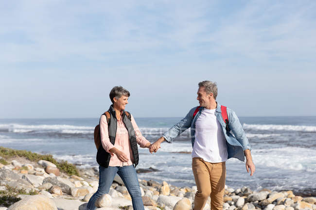 Vista frontal de um casal adulto caucasiano desfrutando de tempo livre andando em uma praia e sorrindo ao lado do mar em um dia ensolarado — Fotografia de Stock