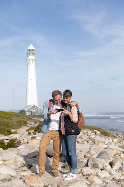 Vorderansicht eines erwachsenen kaukasischen Paares, das an einem sonnigen Tag seine Freizeit mit einem Smartphone genießt und in der Nähe eines Leuchtturms am Meer lächelt — Stockfoto