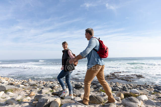 Vista lateral de um casal adulto caucasiano desfrutando de tempo livre andando em uma praia e sorrindo ao lado do mar em um dia ensolarado — Fotografia de Stock