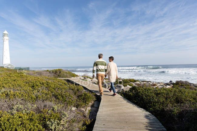 Vista trasera de una pareja caucásica adulta disfrutando de tiempo libre caminando por un sendero junto al mar cerca de un faro en un día soleado - foto de stock