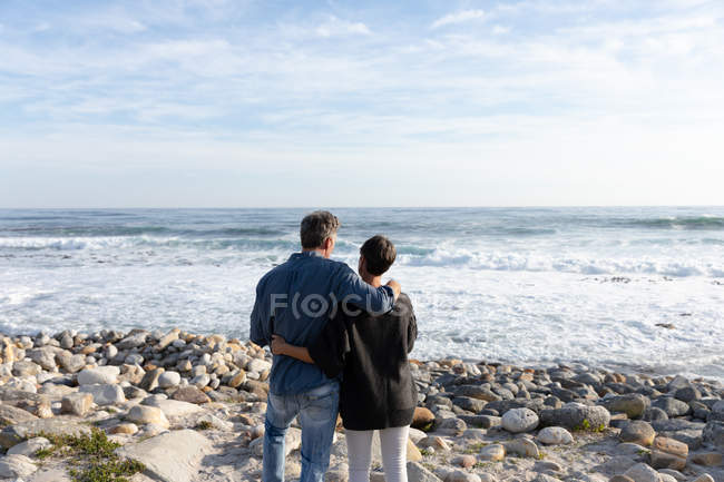 Задний вид на взрослую кавказскую пару, наслаждающуюся свободным временем, обнимающуюся на пляже у моря в солнечный день — стоковое фото