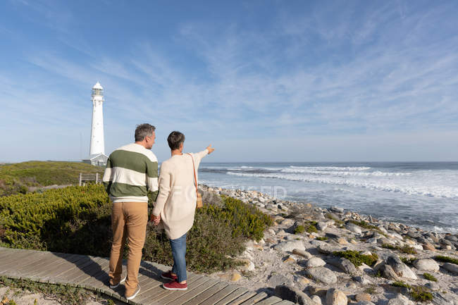 Rear view of an adult Caucasian couple enjoying free time walking a path beside the sea near a lighthouse on a sunny day — Stock Photo