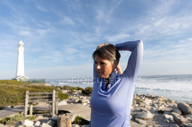 Nahaufnahme einer kaukasischen Frau, die an einem sonnigen Tag in der Nähe eines Leuchtturms am Meer ihre Freizeit beim Stretching genießt — Stockfoto