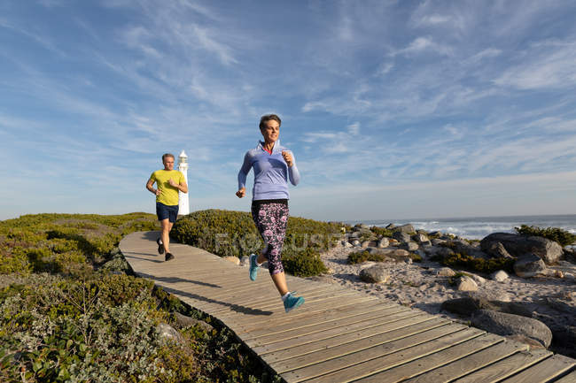 Front view of an adult Caucasian couple enjoying free time running a path near a lighthouse beside the sea on a sunny day — Stock Photo