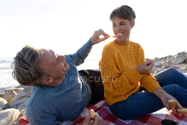 Vista frontale primo piano di una coppia caucasica adulta che si gode il tempo libero rilassandosi insieme su una spiaggia mangiando in una giornata di sole — Foto stock