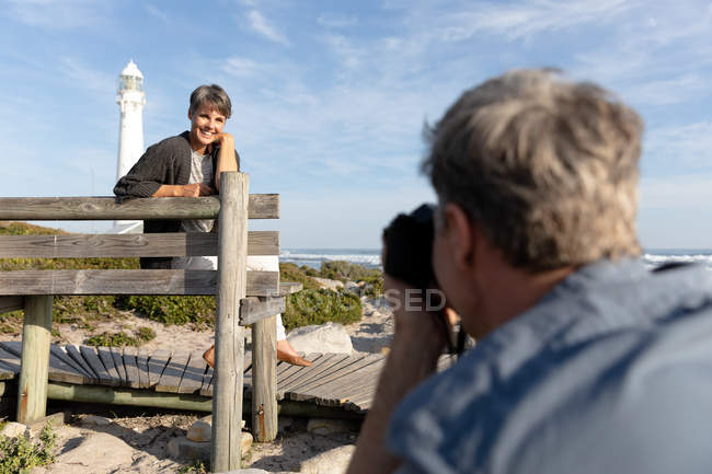 Vista frontal close-up de um casal adulto caucasiano desfrutando de tempo livre tirando uma foto ao lado do mar perto de um farol em um dia ensolarado — Fotografia de Stock