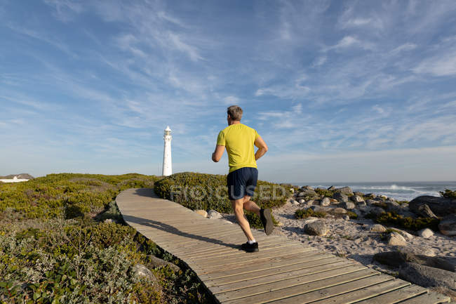 Задний вид на кавказца, наслаждающегося свободным временем, бегущего по тропинке возле маяка у моря в солнечный день — стоковое фото