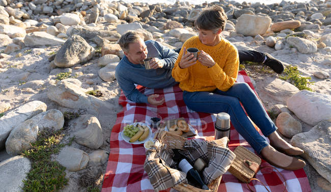 Vorderansicht eines erwachsenen kaukasischen Paares, das an einem sonnigen Tag die freie Zeit am Strand genießt und Kaffee trinkt — Stockfoto