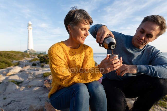 Vista frontale primo piano di una coppia caucasica adulta che si gode il tempo libero rilassandosi insieme su una spiaggia vicino al mare bevendo caffè vicino a un faro in una giornata di sole — Foto stock