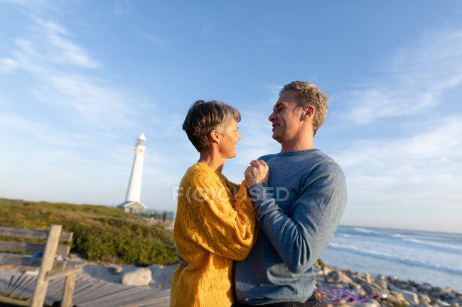 Vista lateral de um casal adulto caucasiano desfrutando de tempo livre relaxando juntos ao lado do mar perto de um farol em um dia ensolarado — Fotografia de Stock