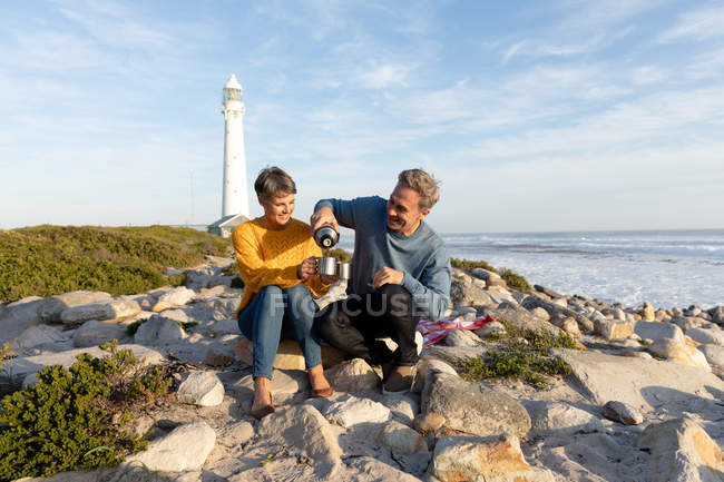 Vista frontale di una coppia caucasica adulta che si gode il tempo libero rilassandosi insieme su una spiaggia vicino al mare bevendo caffè vicino a un faro in una giornata di sole — Foto stock