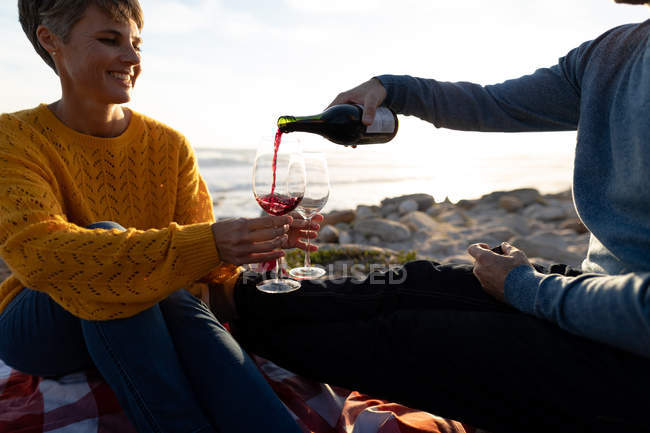 Vista frontal de cerca de una pareja caucásica adulta disfrutando de tiempo libre relajándose juntos en una playa al lado del mar bebiendo vino en un día soleado - foto de stock