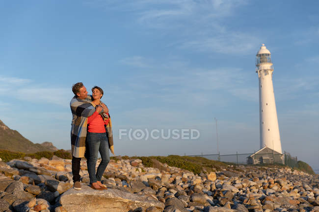 Vue de face d'un couple adulte caucasien jouissant de temps libre debout ensemble embrassant avec une couverture sur leurs épaules près d'un phare par une journée ensoleillée au bord de la mer — Photo de stock