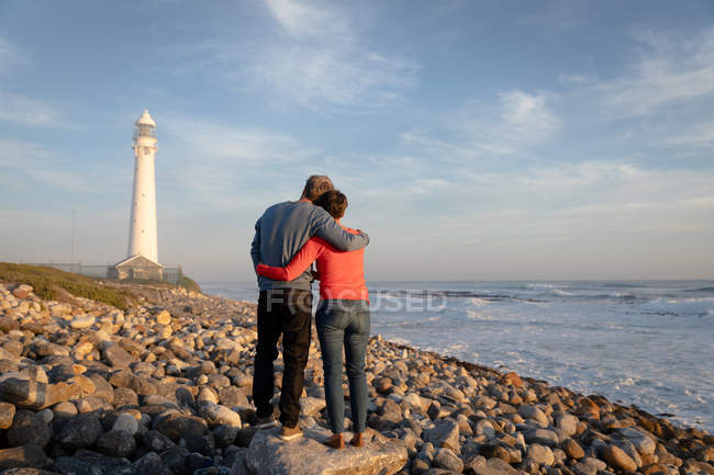 Задний вид на взрослую кавказскую пару, наслаждающуюся свободным временем, расслабляясь вместе, обнимаясь рядом с морем возле маяка в солнечный день — стоковое фото