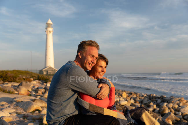 Front view of an adult Caucasian couple enjoying free time relaxing together on a beach embracing together beside the sea near a lighthouse on a sunny day — Stock Photo
