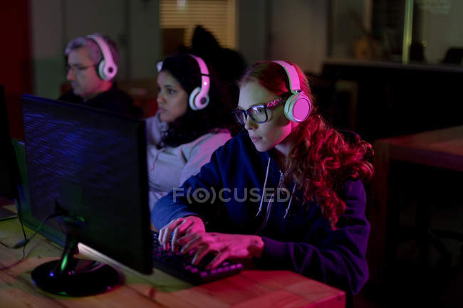 Seitenansicht einer jungen kaukasischen Frau, die in einem kreativen Büro arbeitet, eine Brille trägt, Musik mit Kopfhörern hört und ihre Kollegen im Hintergrund beschäftigt — Stockfoto