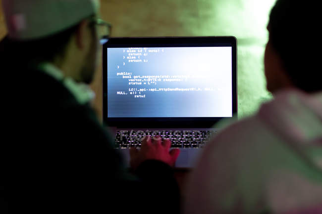 Rückansicht zweier kaukasischer Männer, die in einem kreativen Büro arbeiten, indem sie mit einem Laptop auf den Bildschirm schauen und tippen — Stockfoto