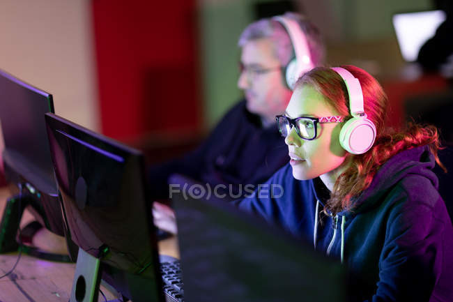 Бічний вид на молоду кавказьку жінку, що працює в творчому офісі, в окулярах, слухаючи музику з навушниками, її колега-чоловік працює на задньому плані — стокове фото