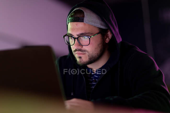 Frontansicht eines jungen kaukasischen Mannes, der in einem kreativen Büro arbeitet, Lesebrille und Kapuze trägt und auf den Computerbildschirm blickt — Stockfoto